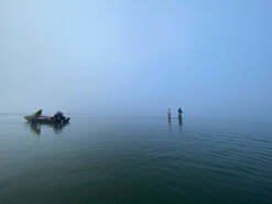 Skiff in the distance with two people standing in knee high water with fog in the background