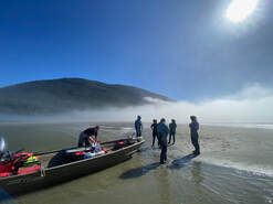 Skiff pulled up on a sandy beach with researchers standing on the beach, fog is clearing off in the background, revealing a mountain and blue sky